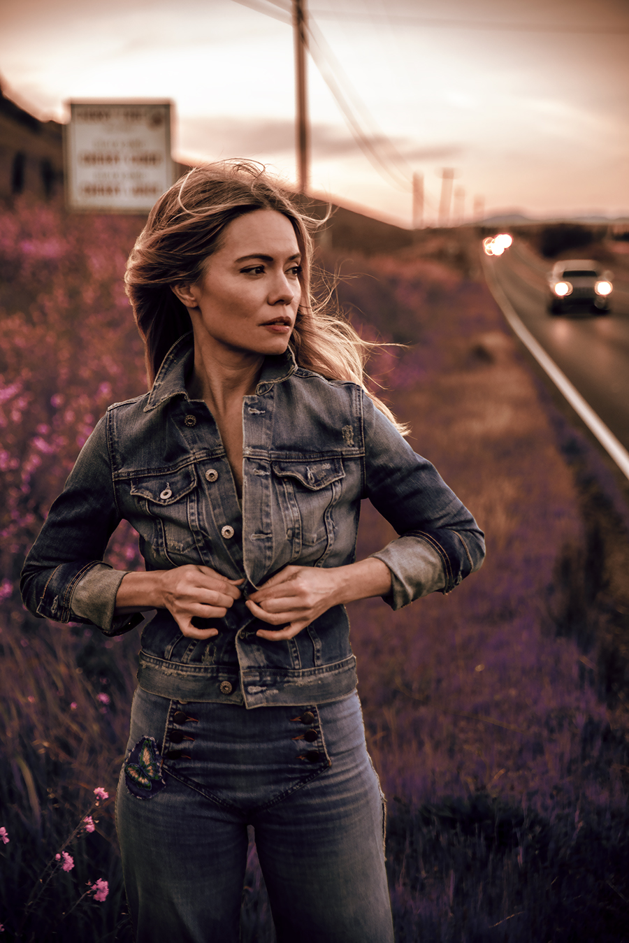 Brooke Geahan stands among the wildflowers by the side of a road. She looks into the distance as the wind blows through her long blonde hair. She is buttoning her denim jacket. The color palette is muted and dramatic.